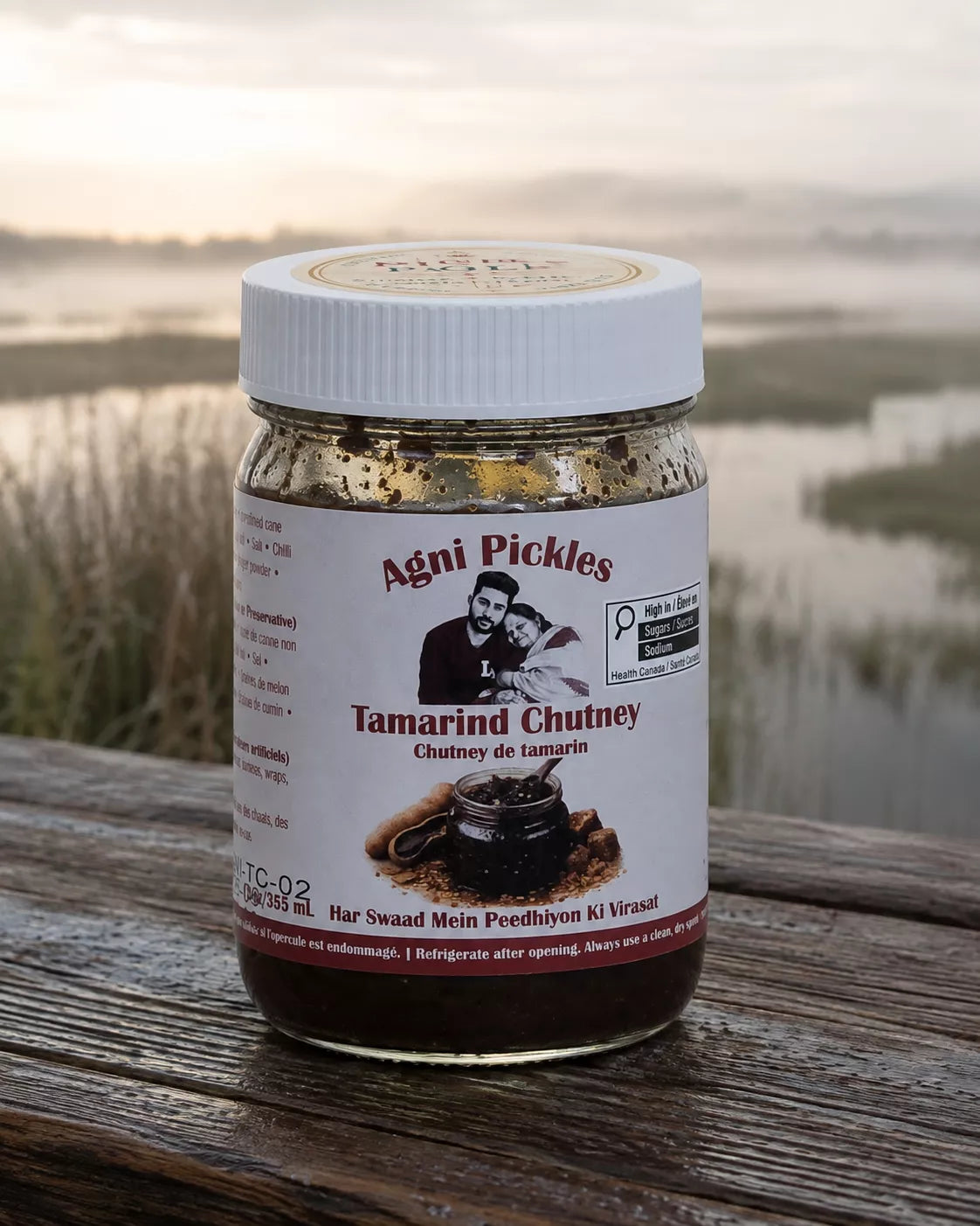 A jar of Agni Pickles Tamarind Chutney sits on a weathered wooden surface with a blurred outdoor background of water and grass at sunset. The jar has a white lid and a white label with red and black text, showing the brand 'Agni Pickles'.