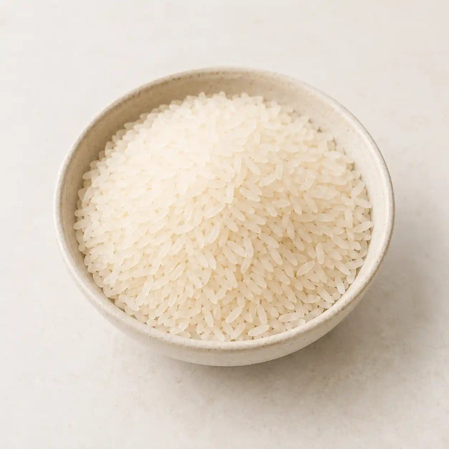 A small beige ceramic bowl filled with uncooked long-grain white rice, photographed from above on a light neutral background.